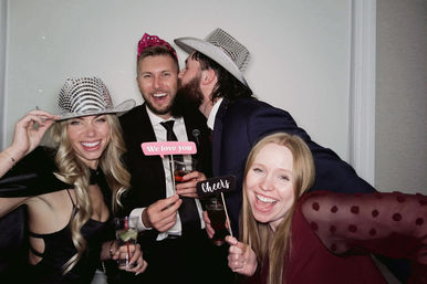 Four friends in a photo booth at an indoor celebration wearing sparkly hats, holding “We love you” and “Cheers” signs; one man kisses another’s cheek as everyone laughs and holds drinks.