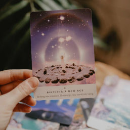 Hand holding a purple cosmic oracle card titled 'Birthing a New Age' depicting a small figure rising in a stone circle under a starry halo