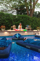 Backyard outdoor pool scene at dusk with people wrapped in blue blankets resting on inflatable mattresses while a facilitator on the deck plays colorful singing bowls and a gong among candles and potted flowers, framed by tall hedges and trees.