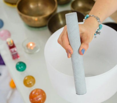 Hand holding a suede mallet over a white crystal singing bowl, surrounded by brass bowls, colorful crystal spheres and a lit tealight — sound healing meditation setup in a wellness studio.