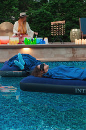 Relaxing outdoor pool scene: two people floating on inflatable mattresses with blankets and an eye mask during a backyard sound-bath surrounded by candles and singing bowls.