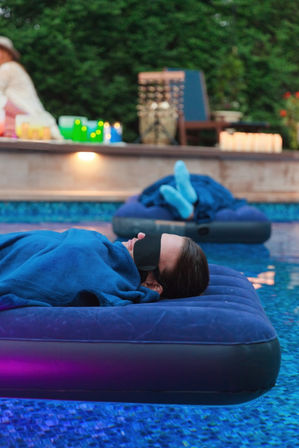 Person lying on a blue inflatable mattress in a backyard pool at dusk, wrapped in a blue blanket and wearing a sleep mask, with a second float, lit candles and soft ambient lighting on the pool deck and mosaic-tile water below.