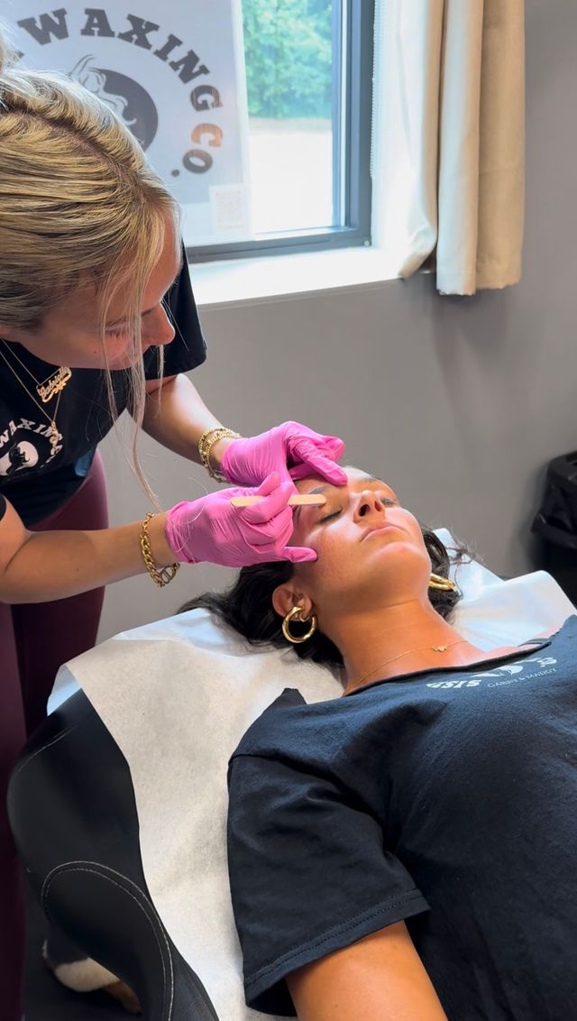 Esthetician wearing pink gloves applying wax with a wooden spatula to a reclining client’s eyebrow during an eyebrow waxing treatment in a beauty studio.