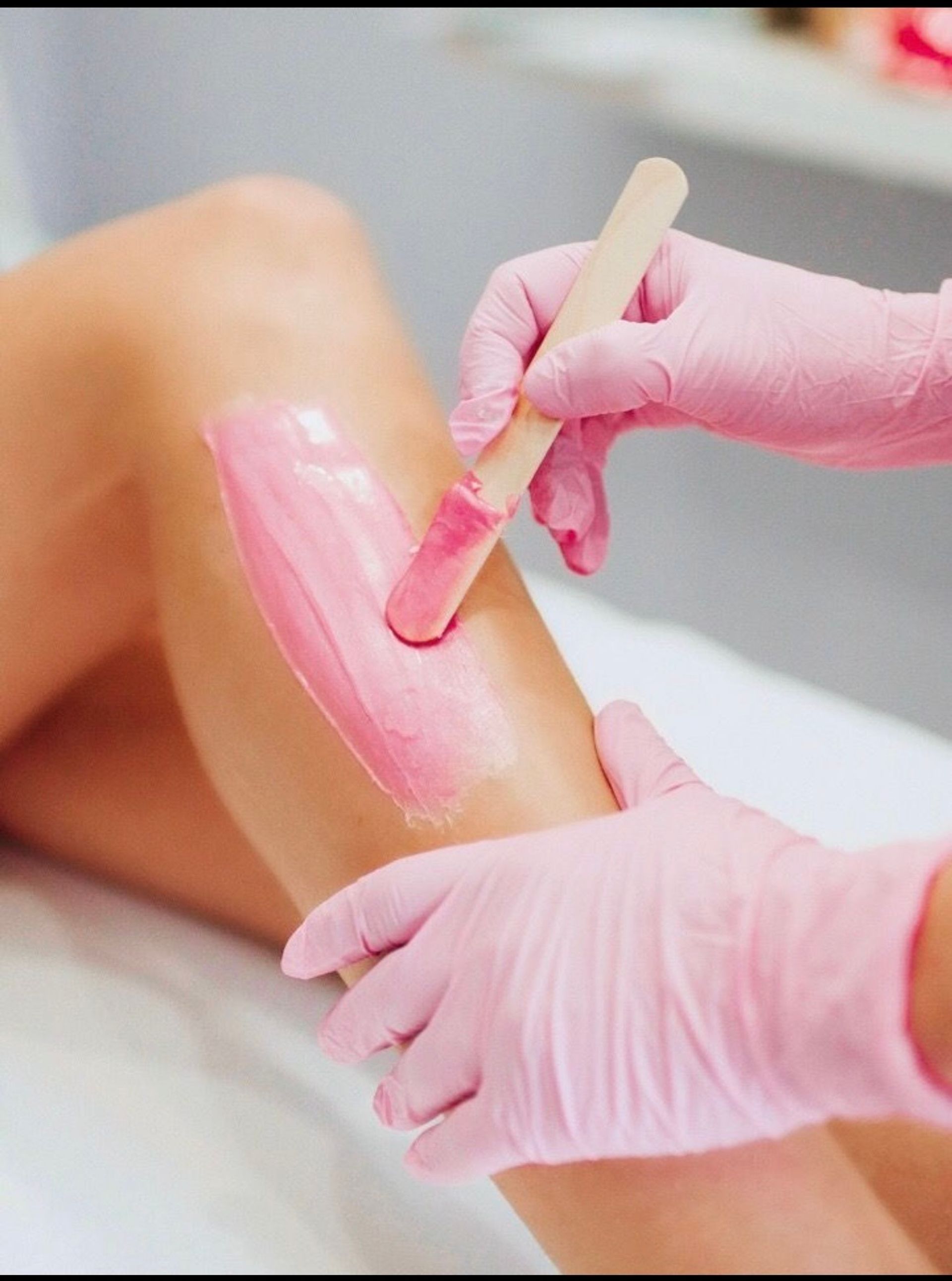 Close-up of leg waxing: technician in pink gloves applying pink wax with a wooden spatula to a lower leg during a spa hair-removal treatment.