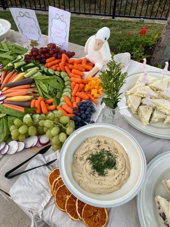 Backyard garden party appetizer spread with colorful crudités (baby carrots, cucumber slices, snap peas, radishes), grapes, blueberries, cheddar cubes, tea sandwiches, a dill-topped dip and dried orange slices, accented by a playful white plush duck.