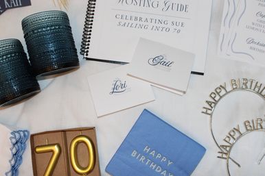 Cheerful flatlay of a 70th birthday party table: blue textured votive holders, gold number candles '70' in a box, blue 'Happy Birthday' napkins, metallic 'Happy Birthday' headbands, nautical-motif place cards and a spiral-bound hosting guide on a white linen surface.