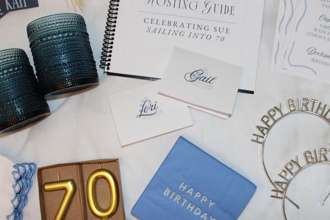 Cheerful flatlay of a 70th birthday party table: blue textured votive holders, gold number candles '70' in a box, blue 'Happy Birthday' napkins, metallic 'Happy Birthday' headbands, nautical-motif place cards and a spiral-bound hosting guide on a white linen surface.