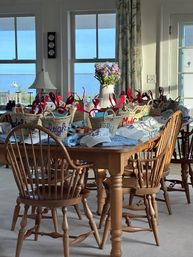 Sunlit coastal dining room in a beach house with a wooden table lined with wicker gift baskets sporting red lobster claws, white caps and goodies, a vase of purple flowers as centerpiece and an ocean view with a boat through the windows