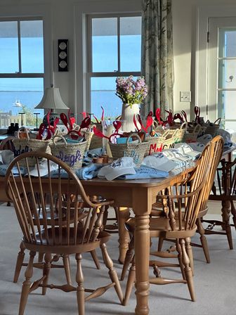 Sunlit seaside dining room with wooden table and chairs, wicker gift baskets topped with red lobster picks and white caps, a vase of purple flowers, and a calm bay with a sailboat visible through the windows.