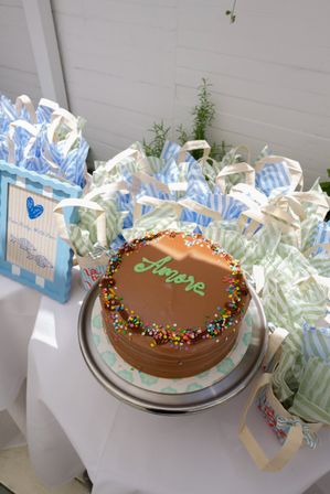 Chocolate frosted cake labeled "Amore" with rainbow sprinkles on a silver stand, surrounded by blue-and-green striped party favor bags on a white dessert table — sunlit garden party dessert setup