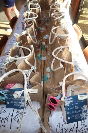 Sunlit rows of burlap beach-style gift bags with sunglasses, teal votive holders and ribbon tags on a rustic wooden table — coastal party favors in Carmel
