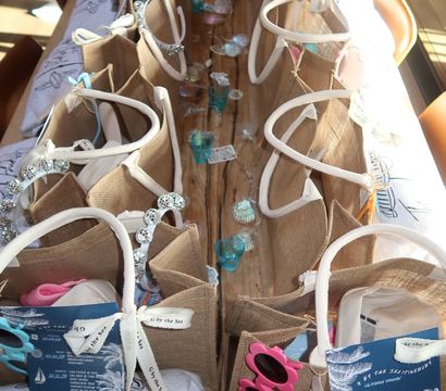 Sunlit rows of burlap beach-style gift bags with sunglasses, teal votive holders and ribbon tags on a rustic wooden table — coastal party favors in Carmel