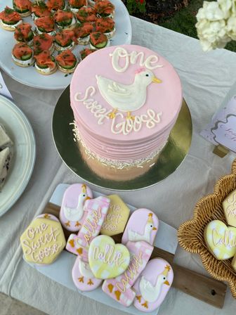 Pink two-layer birthday cake topped with a white goose and “one sweet goose” script, surrounded by matching goose-themed sugar cookies and savory canapés on an outdoor dessert table.