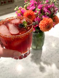 Hand holding a bright red spicy cocktail in a short rocks glass with a chili-salt rim, ice cubes, blackberry and jalapeño garnish on a pick, blurred orange and pink flowers in a vase on a marble countertop.