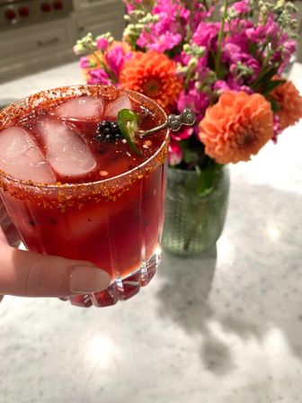Hand holding a spicy red berry cocktail in a rocks glass with ice, chili-salt rim and jalapeño garnish on a marble kitchen countertop with bright orange and pink flowers in a vase.