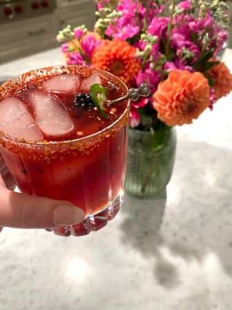 Close-up of a vibrant red spicy berry cocktail in a faceted glass with chili-salt rim, ice, blackberry and jalapeño garnish, held above a marble countertop with bright orange and pink flowers in a vase.
