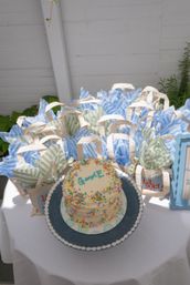 Small two-tier white cake with multicolored sprinkles and a handwritten message on a blue platter, surrounded by blue- and green-striped tissue-filled tote favor bags on a white table at an outdoor party