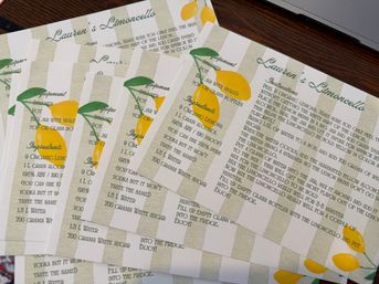 Stack of printable limoncello recipe cards with bright lemon illustrations and striped background, fanned out on a wooden table next to a laptop