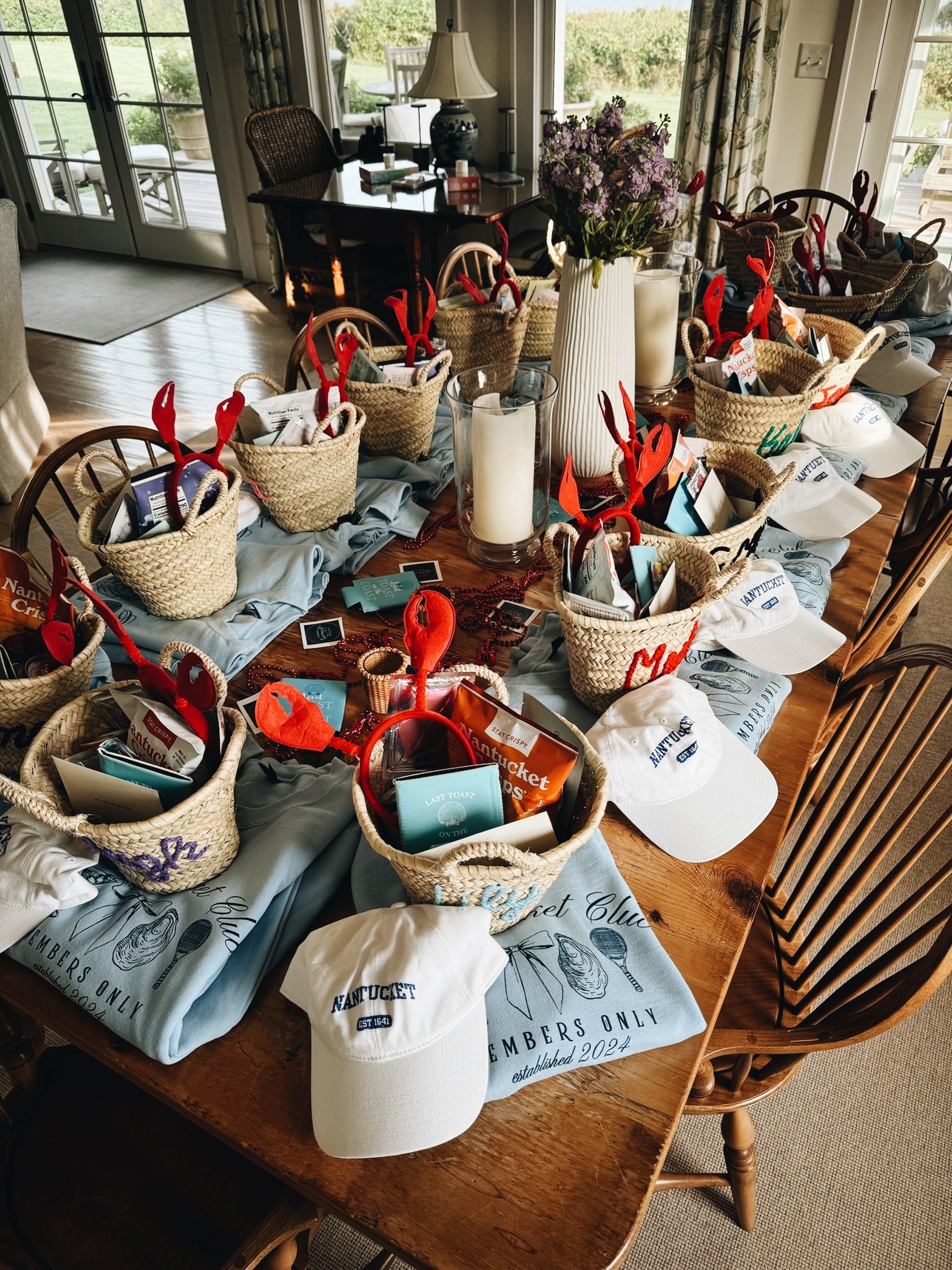 Sunlit wooden dining table in a Nantucket beach house set with woven gift baskets of snacks, red lobster-claw headbands, white ‘Nantucket’ caps, folded blue club tees, candles, and a vase of purple flowers — festive seaside welcome gifts.