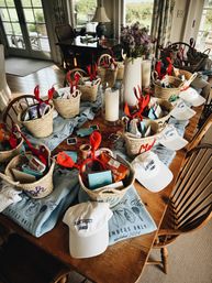 Nantucket-themed welcome spread on a wooden dining table: woven straw baskets filled with snacks, cards and red lobster headbands, white "Nantucket" caps and light-blue "Members Only" shirts, with a vase of purple flowers and candles.