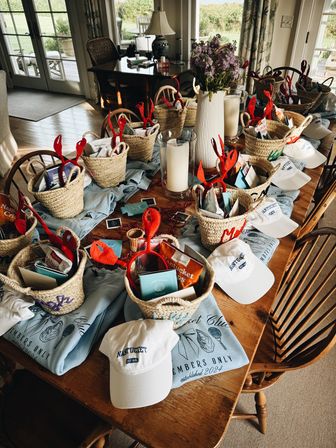 Sunlit wooden dining table in a Nantucket beach house set with woven gift baskets of snacks, red lobster-claw headbands, white ‘Nantucket’ caps, folded blue club tees, candles, and a vase of purple flowers — festive seaside welcome gifts.