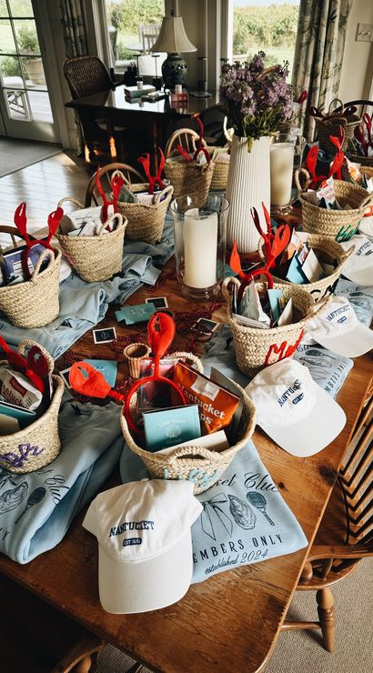 Sunlit wooden dining table in a Nantucket beach house set with woven gift baskets of snacks, red lobster-claw headbands, white ‘Nantucket’ caps, folded blue club tees, candles, and a vase of purple flowers — festive seaside welcome gifts.