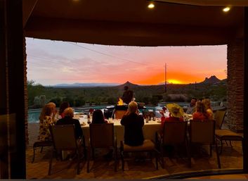 Guests enjoying an al fresco dinner under a covered patio while a chef grills by a pool, framed by a vibrant orange Southwestern desert sunset and silhouetted mountains.