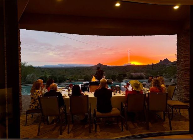 Guests enjoying an al fresco dinner under a covered patio while a chef grills by a pool, framed by a vibrant orange Southwestern desert sunset and silhouetted mountains.