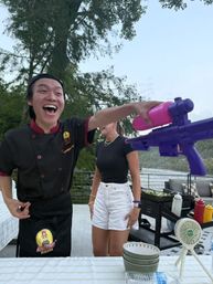 Laughing chef in black uniform reaches toward a purple water gun as a woman in a black top and white shorts stands on a lakeside deck with grill, condiments and bowls during a summer outdoor gathering