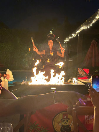 Backyard poolside fire performer at night juggling flaming torches above a rectangular fire pit, outdoor string lights and guests filming the fiery show with their phones