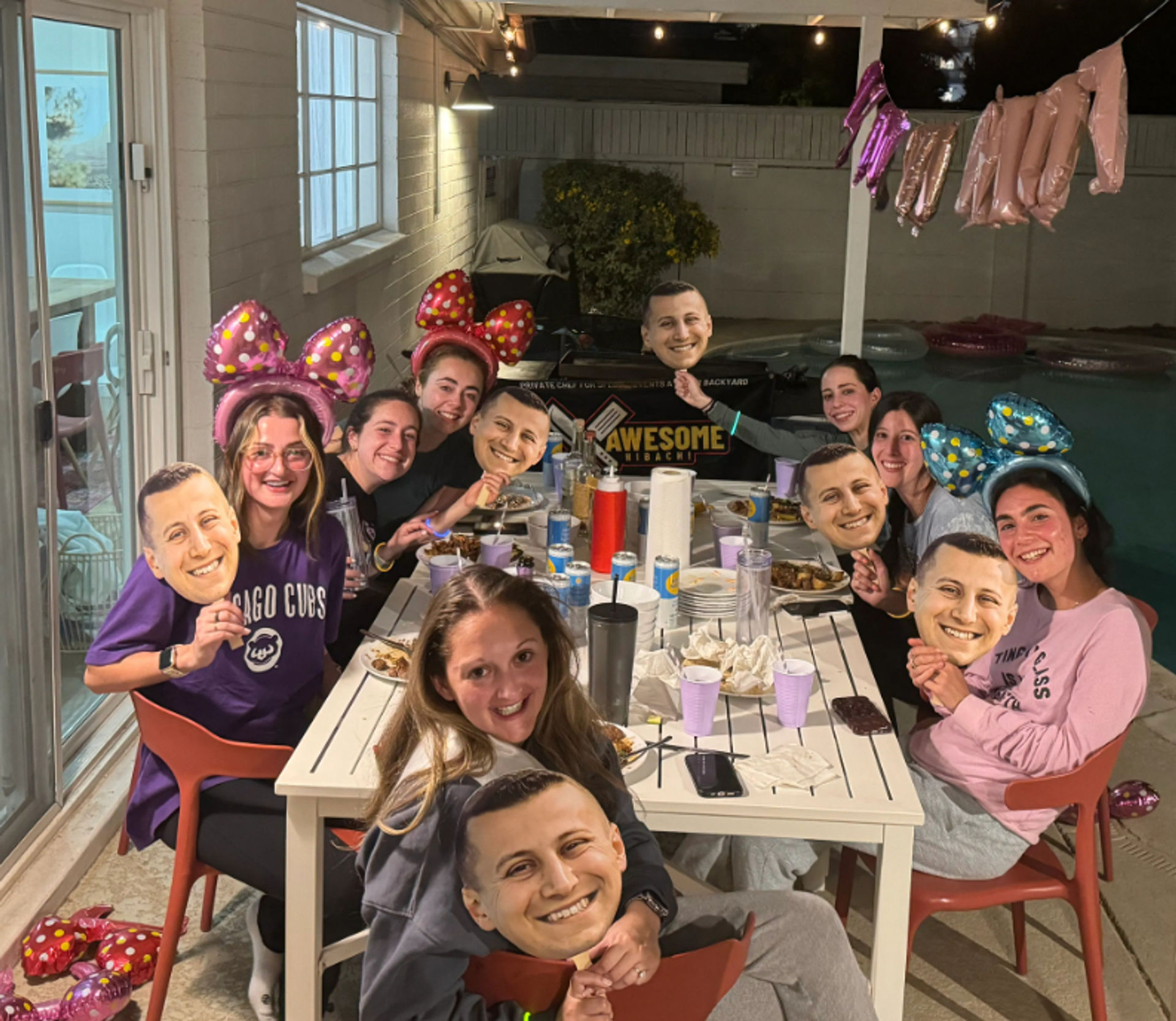 Backyard poolside patio dinner party at night with a group of friends around a table, wearing polka-dot bow headbands and holding identical cardboard face masks, plates and cups on the table, string lights and pink foil balloons overhead.