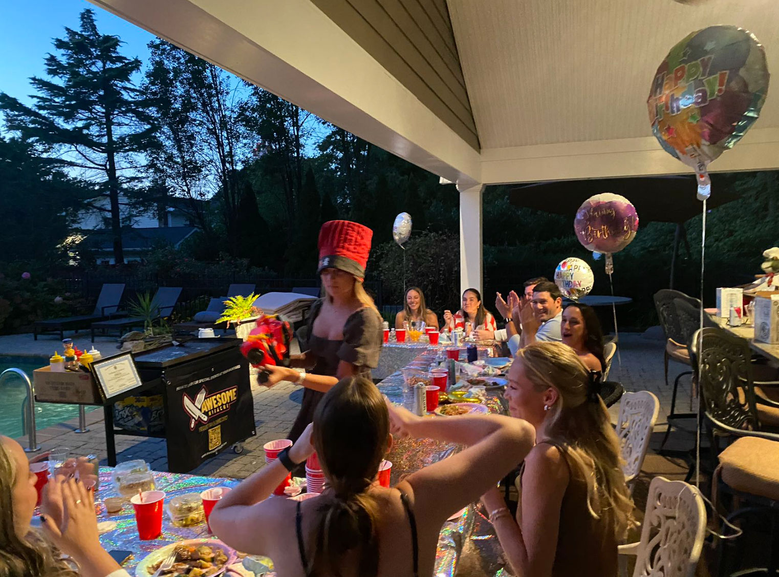 Backyard poolside birthday party on a covered patio at dusk — friends seated along a long table with red cups, plates and balloons while a woman in a tall red novelty hat presents a gift.