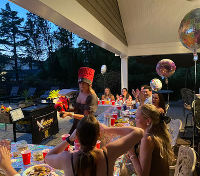 Backyard poolside birthday party on a covered patio at dusk — friends seated along a long table with red cups, plates and balloons while a woman in a tall red novelty hat presents a gift.