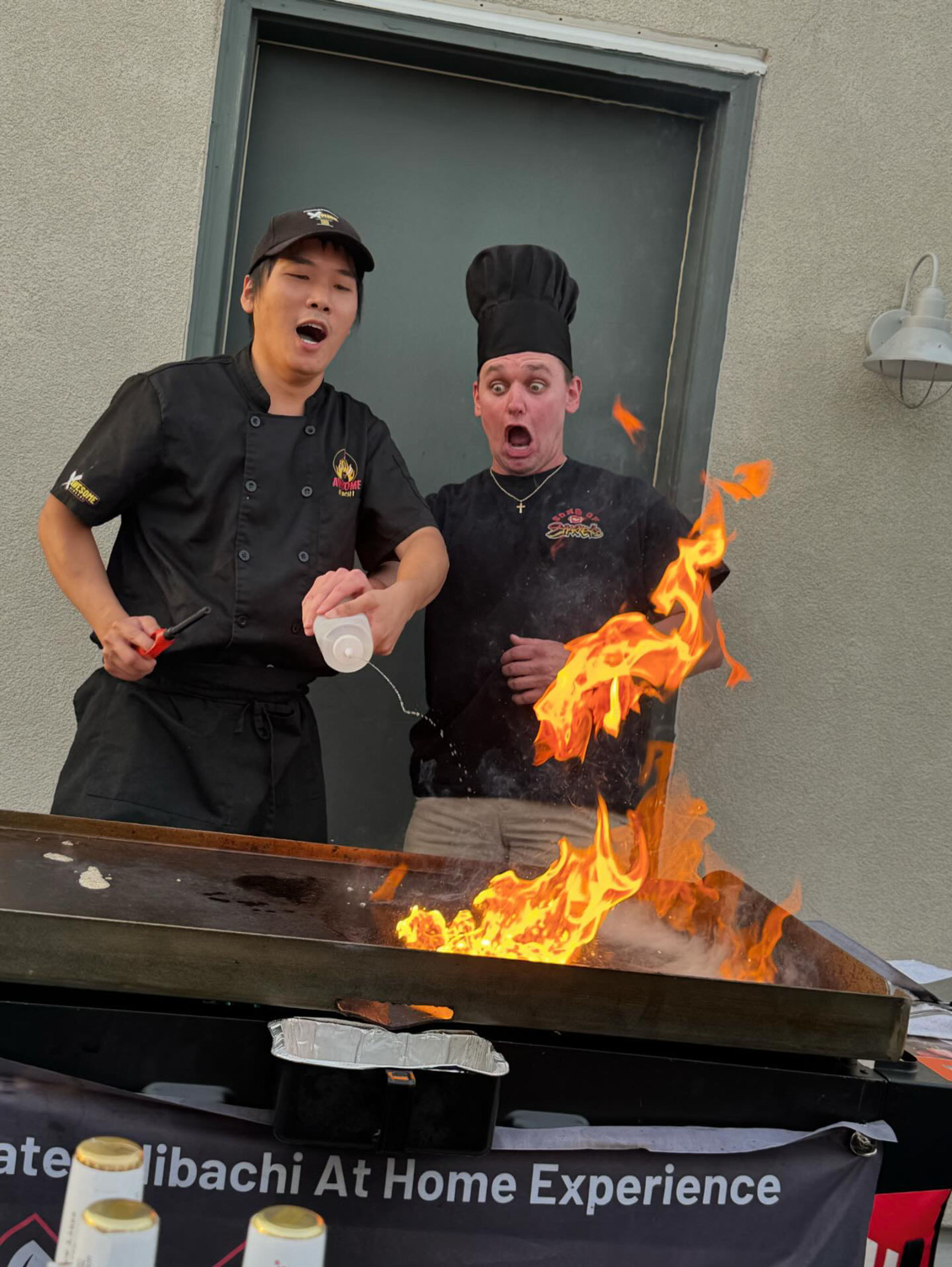 Two chefs at an outdoor teppanyaki griddle react with surprised expressions as a large orange flame erupts when one pours liquid onto the hot surface.