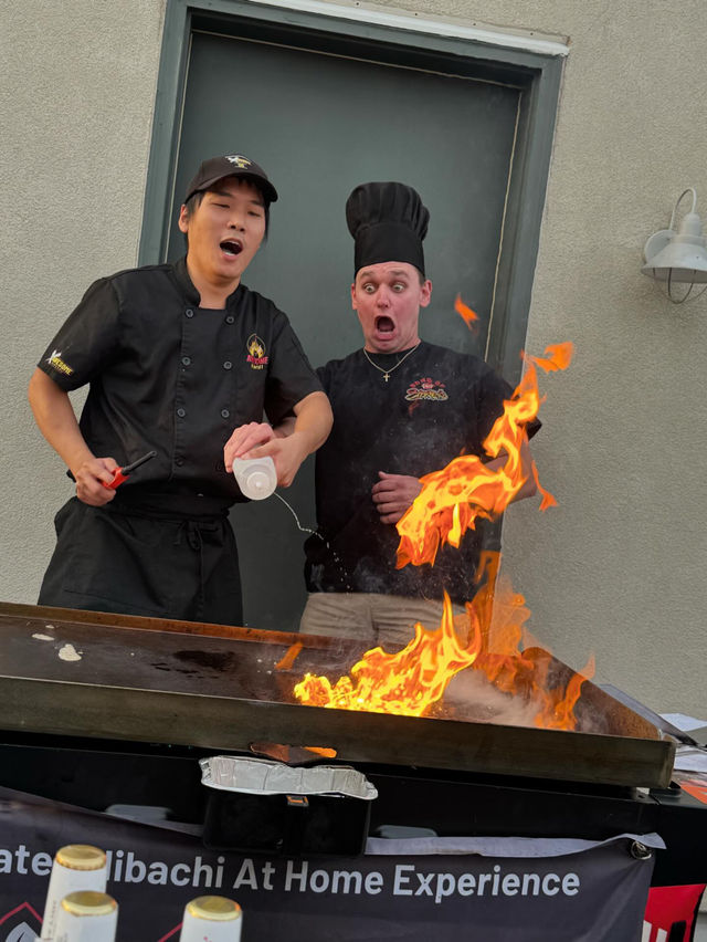 Two chefs at an outdoor teppanyaki griddle react with surprised expressions as a large orange flame erupts when one pours liquid onto the hot surface.