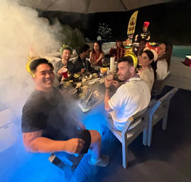 Backyard poolside party at night with friends gathered around a long outdoor patio table, holding drinks and wearing leis and festive hats, plates of food, blue pool lights and party smoke creating a lively atmosphere.