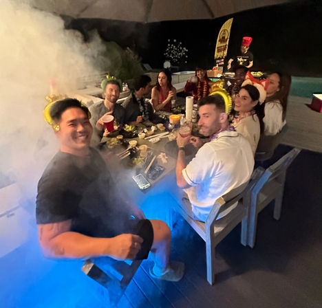 Backyard poolside party at night with friends gathered around a long outdoor patio table, holding drinks and wearing leis and festive hats, plates of food, blue pool lights and party smoke creating a lively atmosphere.