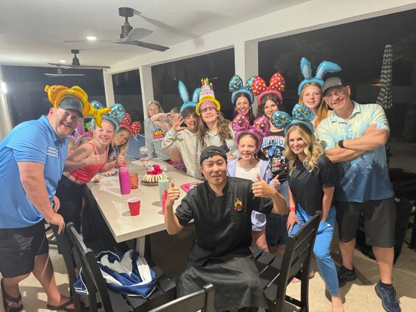 Group birthday celebration on a covered backyard patio at night, guests wearing colorful balloon ears and bunny hats around a table with a cake, a person in black uniform giving thumbs up — festive scene.