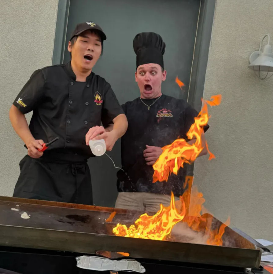 Two chefs at an outdoor flat-top grill react in shock as a pour sparks a dramatic flame burst during a teppanyaki-style cooking demo