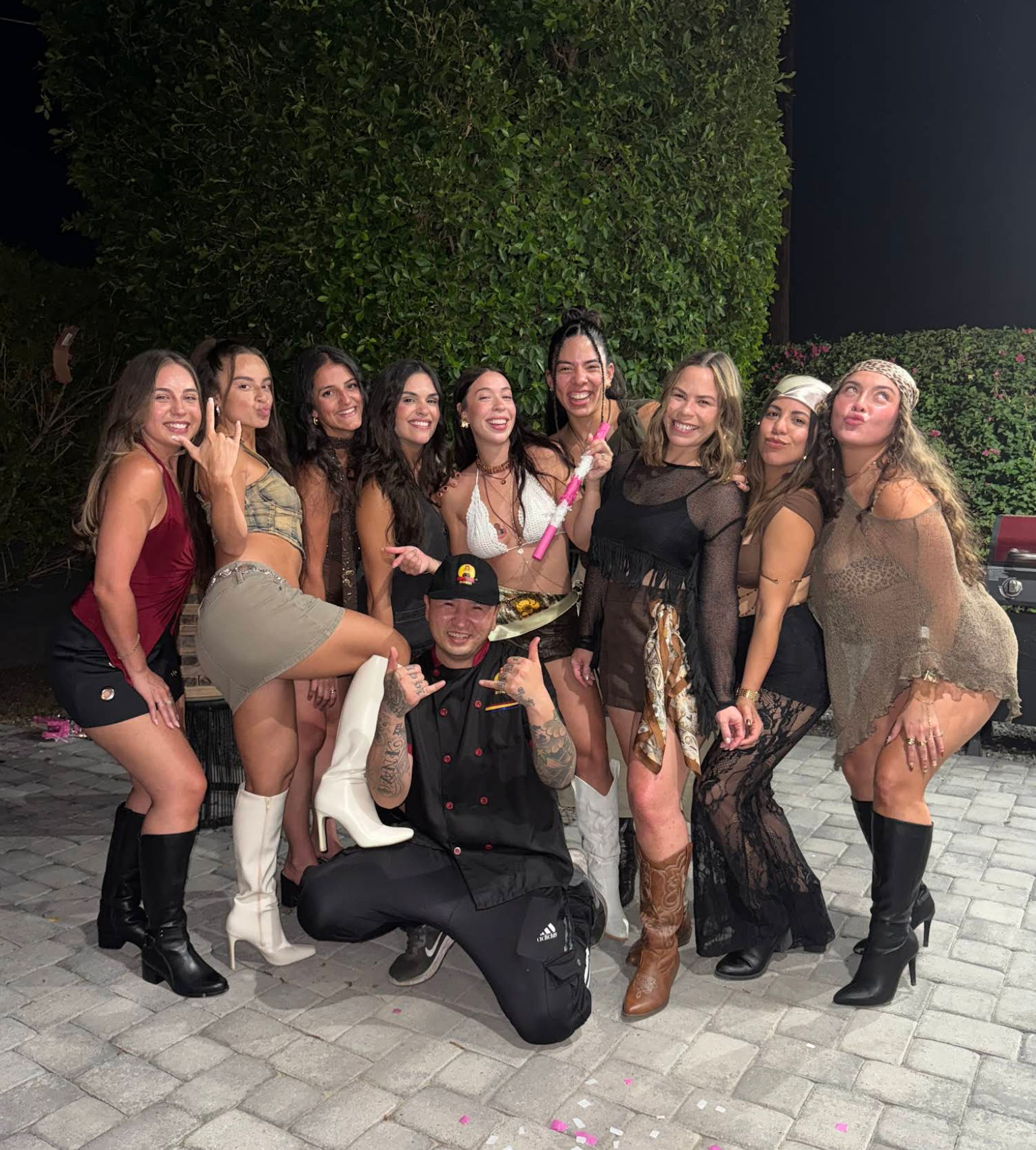 Ten friends posing and laughing in stylish party outfits and boots on a paved backyard patio at night with a tall hedge backdrop