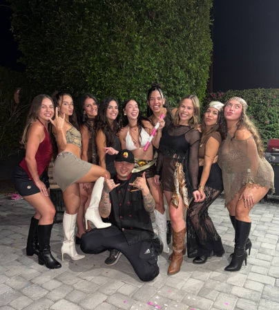 Ten friends posing and laughing in stylish party outfits and boots on a paved backyard patio at night with a tall hedge backdrop