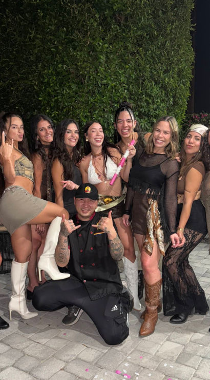 Ten friends posing and laughing in stylish party outfits and boots on a paved backyard patio at night with a tall hedge backdrop