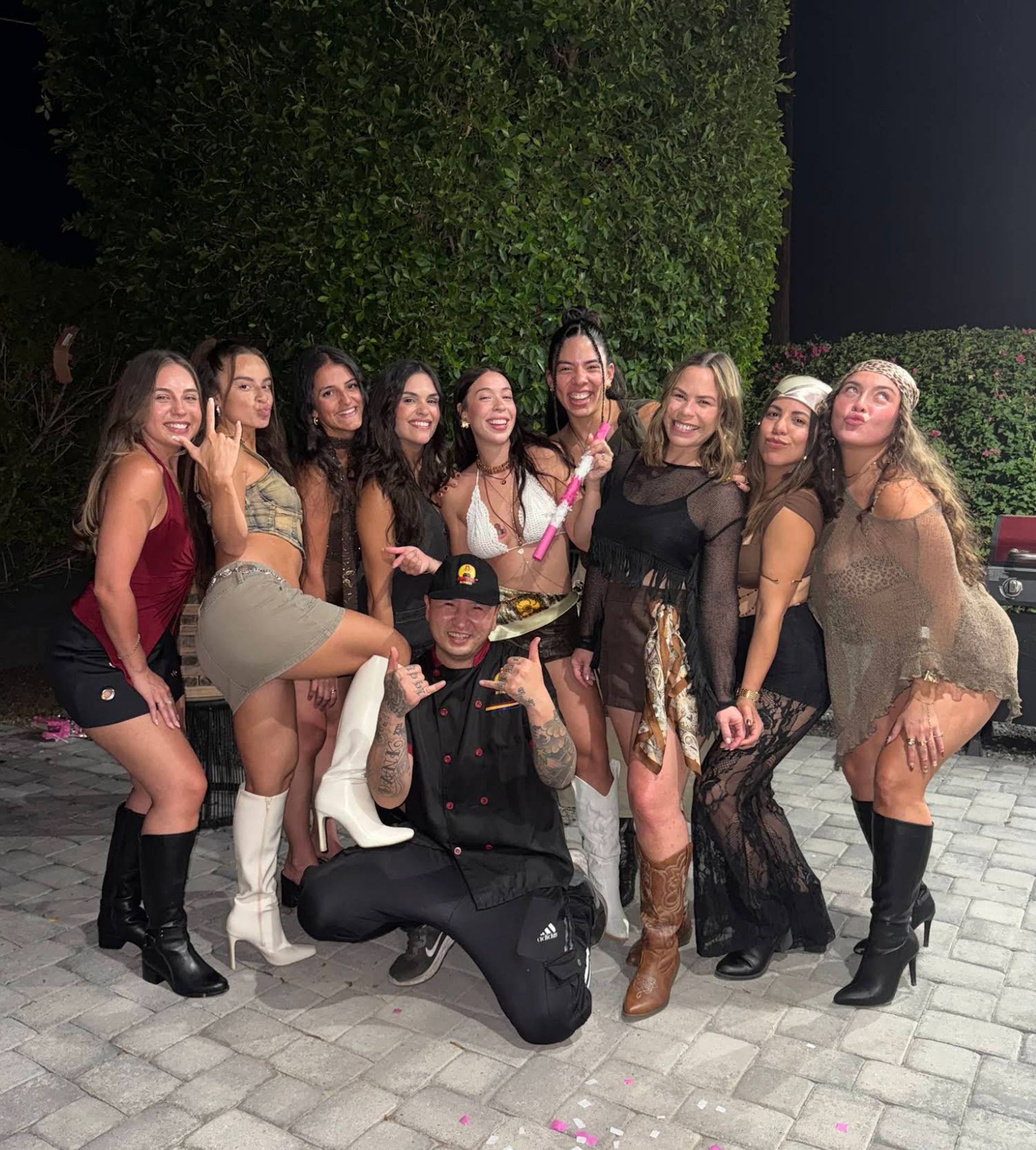Ten friends posing and laughing in stylish party outfits and boots on a paved backyard patio at night with a tall hedge backdrop