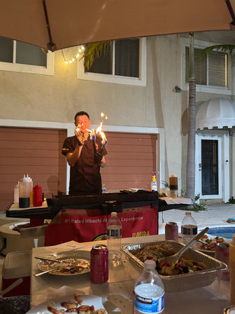 Poolside hibachi chef performing a fiery griddle show on an outdoor patio at a backyard dinner, flames rising over the grill with foil trays of rice and vegetables and drinks on the table.