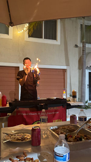 Poolside hibachi chef performing a fiery griddle show on an outdoor patio at a backyard dinner, flames rising over the grill with foil trays of rice and vegetables and drinks on the table.