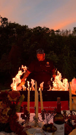 Outdoor sunset scene of a chef theatrically cooking over tall flames behind a candlelit table set with wine, flowers and olives.