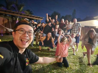 Lively backyard group selfie at night: smiling adults and kids posing on a grassy lawn under evening lights.