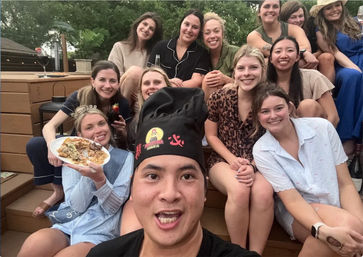 Smiling group selfie on a backyard wooden deck: friends gathered around a man in a chef hat with a plate of food and drinks at a casual outdoor party.
