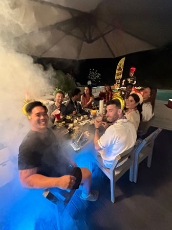 Nighttime backyard poolside party with friends gathered around a table of food and drinks under a large umbrella, wearing colorful hats and leis, with smoky blue lighting and a server or chef in the background.