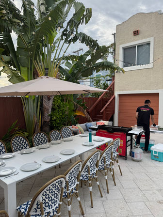Tropical backyard patio set for outdoor dining — long white table with plates and blue-patterned chairs under an umbrella, string lights overhead, banana palms, and a cook prepping at a portable grill beside a stucco house and garage.